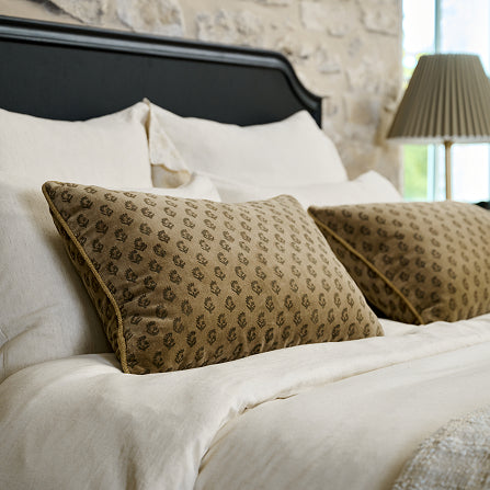 Close-up of a bed with patterned pillows and a lamp in the background.  Shop neutral bedding.
