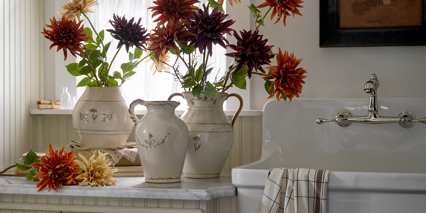 Decorative vases with autumn flowers on a marble countertop next to a white sink.