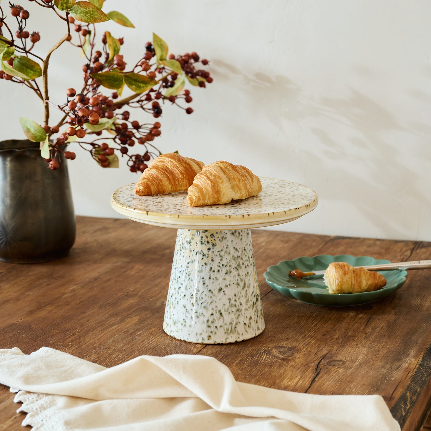 Crescent rolls on a decorative stand with a vase of berries and a plate of croissant on a wooden table.