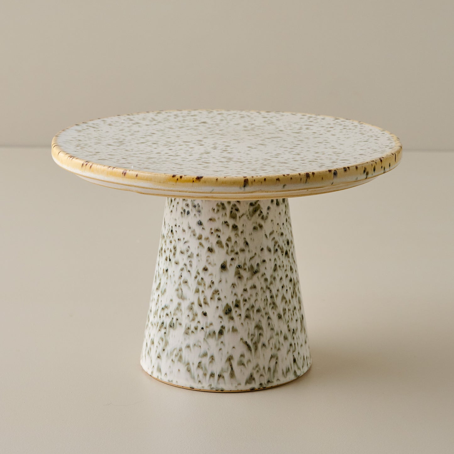 Green speckled ceramic cake stand with a round top and cylindrical base on a beige background