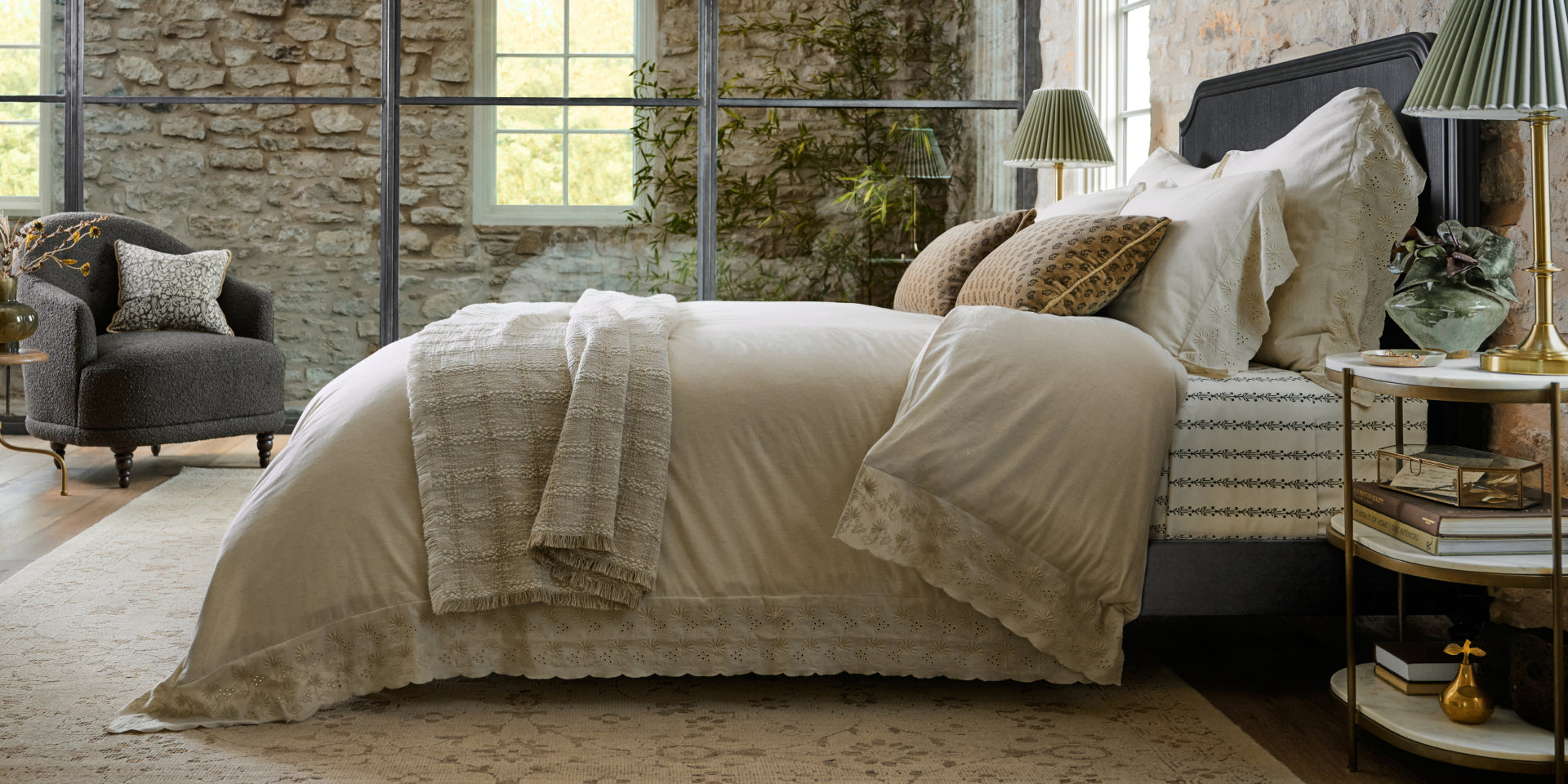 Neatly made bed in a room with stone walls and large windows. Shop all Bedding.