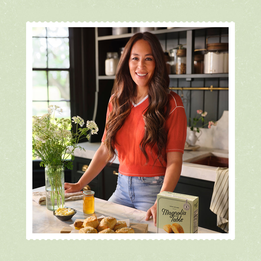 Joanna Gaines in her butler's pantry with a box of Frozen Magnolia Table buttermilk biscuits along with a wooden tray full of baked Magnolia Table buttermilk biscuits.