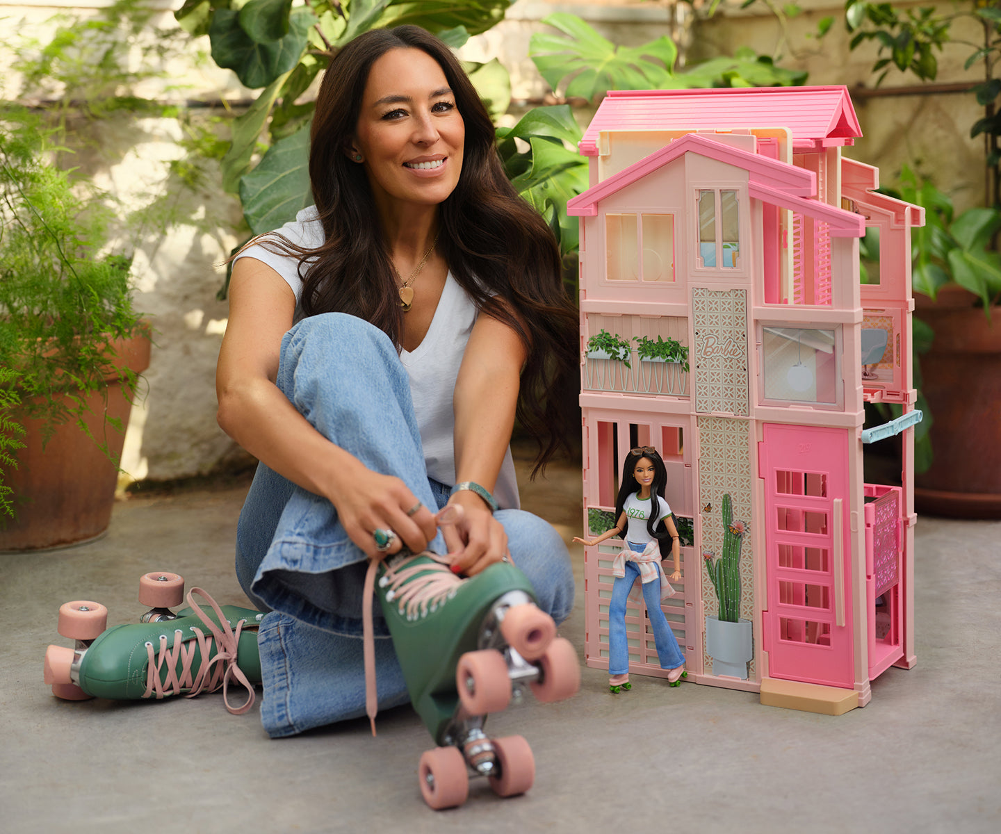 Joanna Gaines sitting on the ground with a pink dollhouse and Barbie doll in an outdoor setting