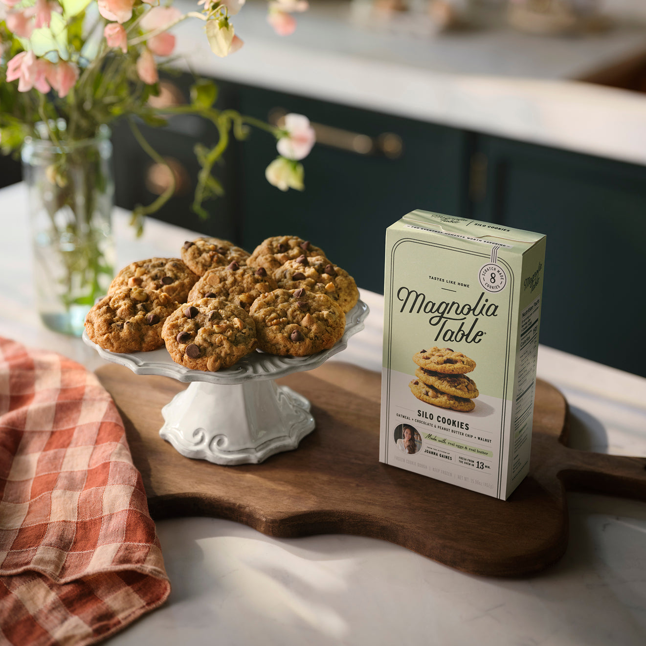 Box of Magnolia Table Frozen Silo Cookie Dough on a kitchen island with Magnolia Table Silo Cookies on a plate stand and flowers in the background.