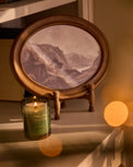 An oval framed landscape print on a small wooden stand, styled on a shelf next to a lit green candle with warm bokeh lights in the background.