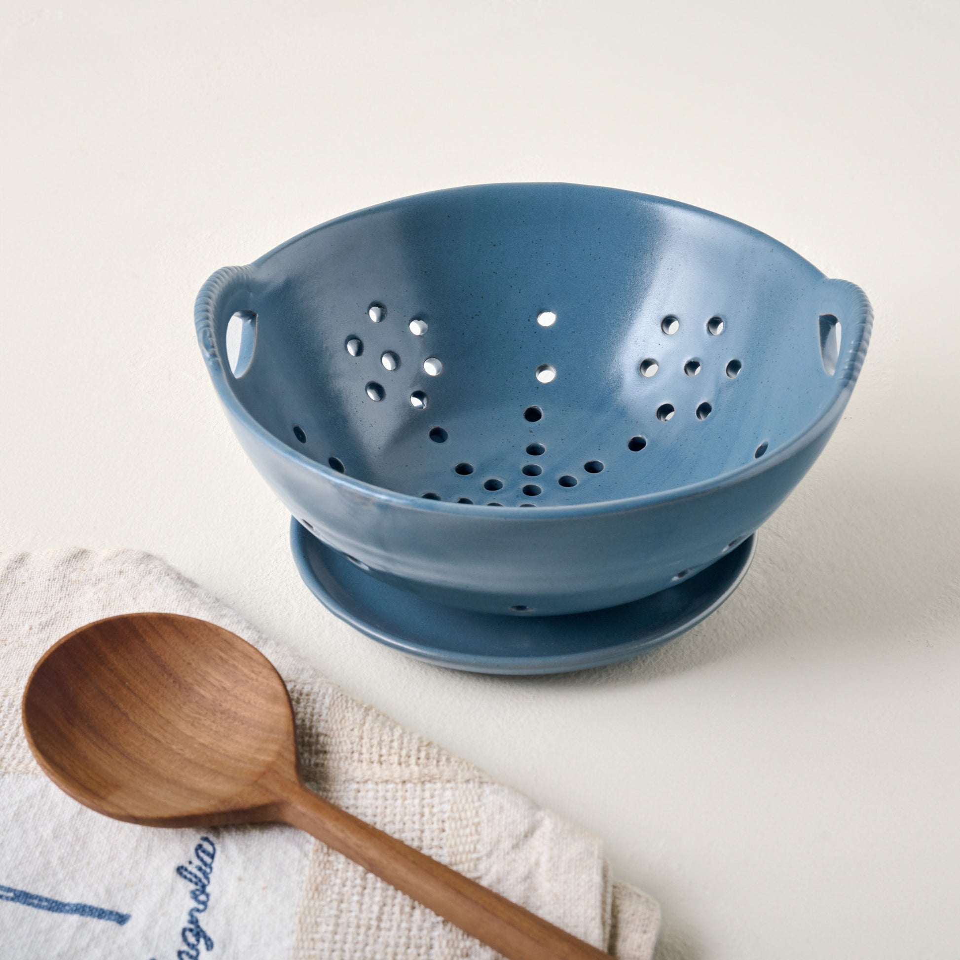 Maribel Stoneware Colander + Plate shown with tea towel and wooden spoon
