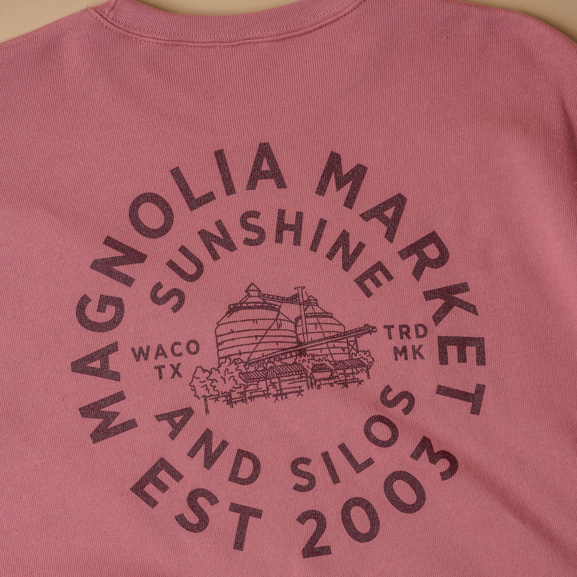 Close up of back of mauve colored sweatshirt in a circular shape reads Magnolia Market Est 2003. There is an inner circle of text that reads sunshine and silos. In the center of the circle of text is a picture of the silos and on either side of the picture reads Waco TX and TRD MK. Sweatshirt on model with back turned. All text is in maroon color.