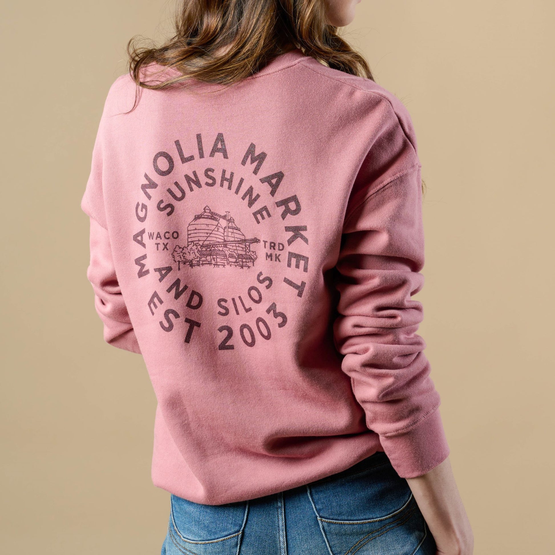 Back of mauve colored sweatshirt in a circular shape reads Magnolia Market Est 2003. There is an inner circle of text that reads sunshine and silos. In the center of the circle of text is a picture of the silos and on either side of the picture reads Waco TX and TRD MK. Sweatshirt on model with back turned.