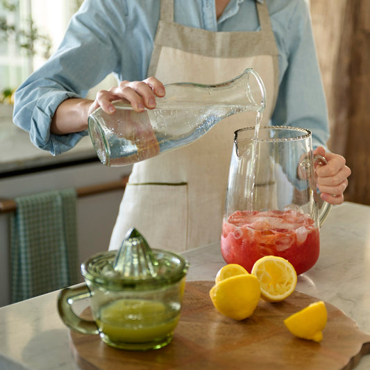 A woman making sparkling lemonade with a glass juicer, fresh lemons, and a large pitcher of strawberry puree.