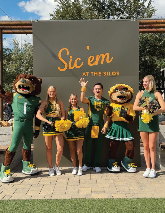 The Baylor Bears mascots with the Baylor cheerleaders in front of a green banner that says Sic'em at the Silos.