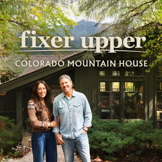 A square image of Chip and Joanna Gaines standing in front of the home featured on Fixer Upper: Colorado Mountain House—surrounded by lush greenery and landscape.