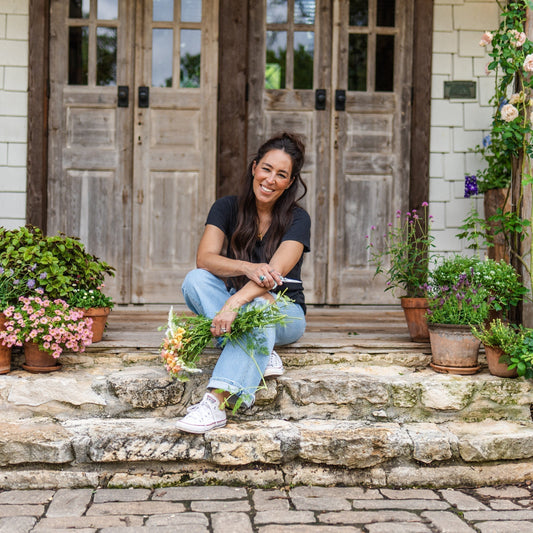 Joanna Gaines smiles on her front porch—surrounded by plants and holding a bundle of fresh flowers.
