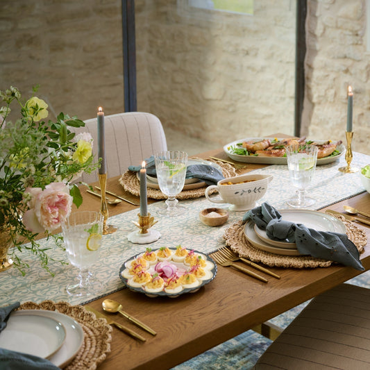 A wooden dinner table dressed for Easter with woven placemats, candles, and delicious dishes like deviled eggs.