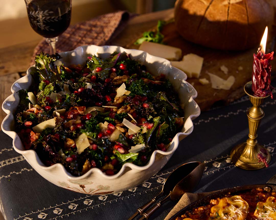 A large wooden bowl of kale salad tossed with crispy prosciutto, candied walnuts, pomegranate, and Parmesan cheese near a lit taper candle.