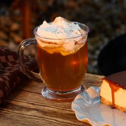 A mug of maple apple cider and a slice of Candy Bar cheesecake on a small wooden table near a fire pit.