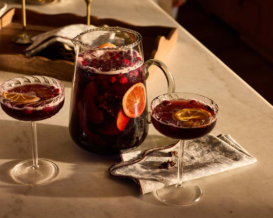 A pitcher and two glasses of a red wine sangria filled with cranberries and orange slices.