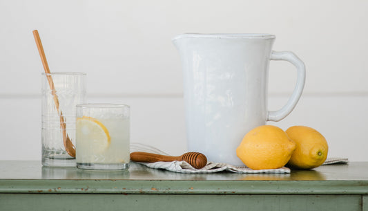 A white pitcher, kitchen tools, and a glass of lemonade sit on a green tabletop. 