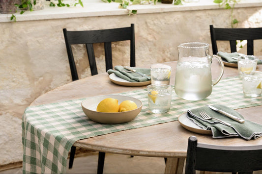 A bowl of two yellow lemons sits on a green gingham table runner. Plates, topped with green table napkins, and glassware sit on a wooden table from Magnolia.