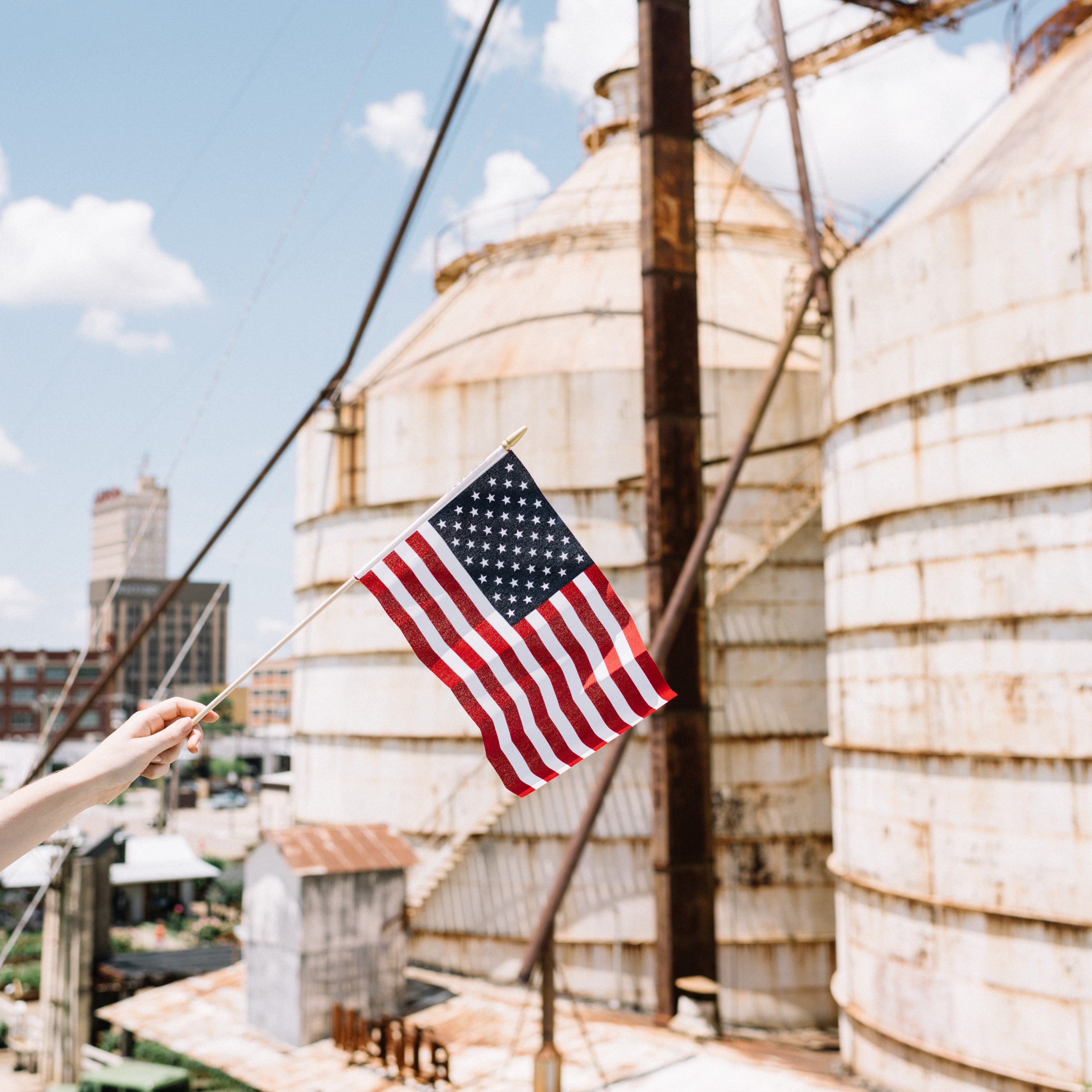American flag held by a person in front of large industrial tanks under a blue sky.