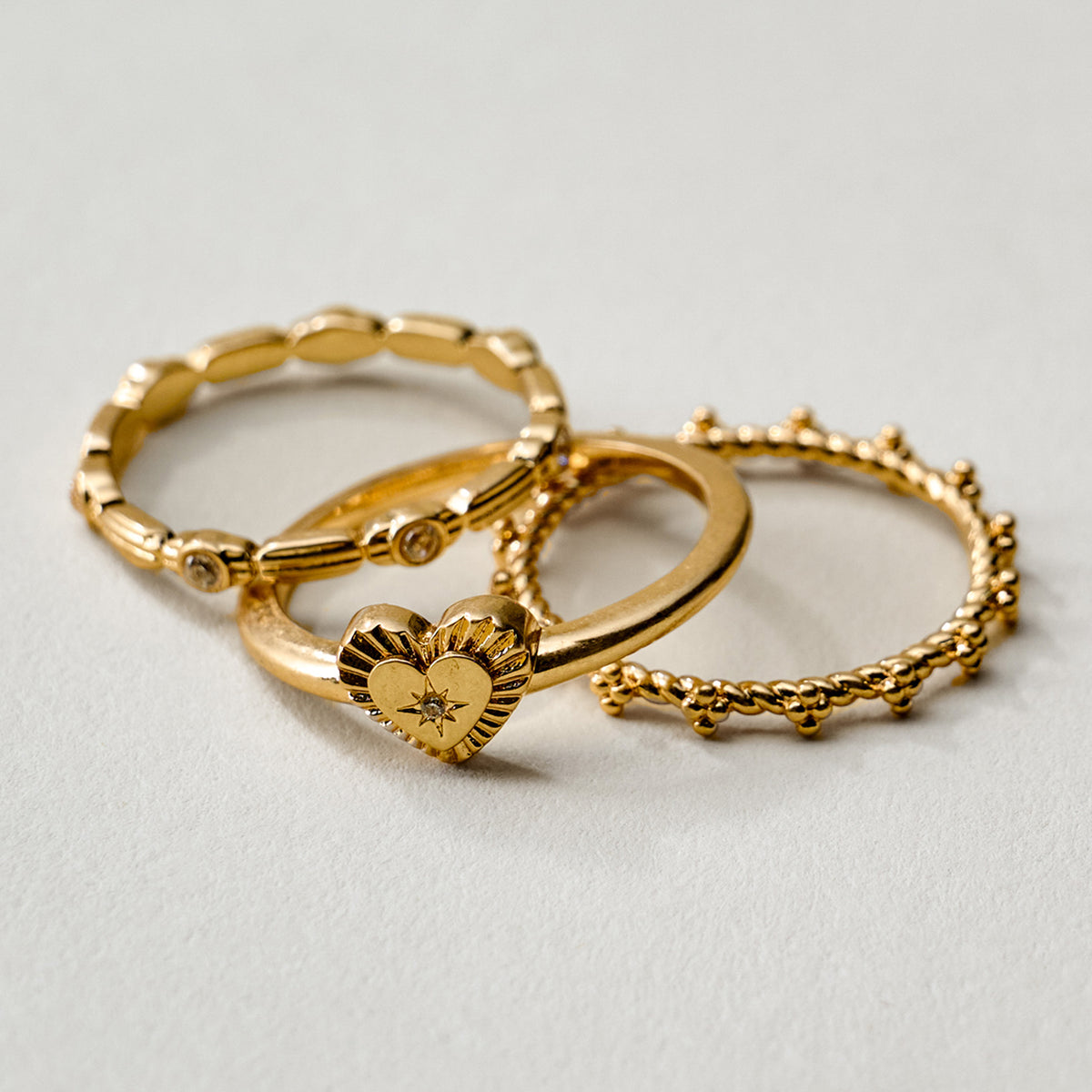 Three gold rings with textured and sculpted designs, including a heart-shaped ring with a small clear gemstone, arranged on a light gray background.