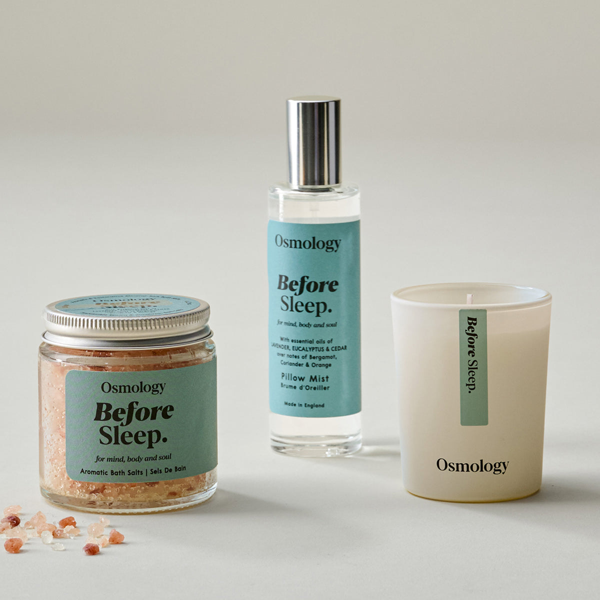 Three Osmology 'Before Sleep.' products arranged on a light neutral background: an aromatic bath salt jar with pink crystals, a clear bottle of pillow mist with a silver cap, and a cream candle in a white vessel.