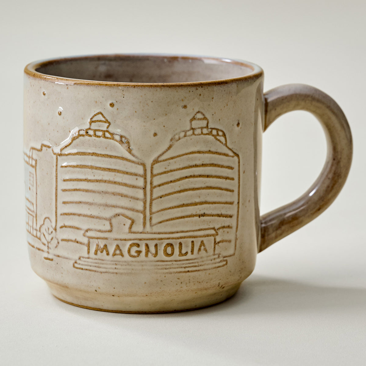 A handmade ceramic coffee mug with a speckled beige and brown glaze, featuring an embossed design of two magnolia trees and the word 'MAGNOLIA' on the front, set against a plain light background.