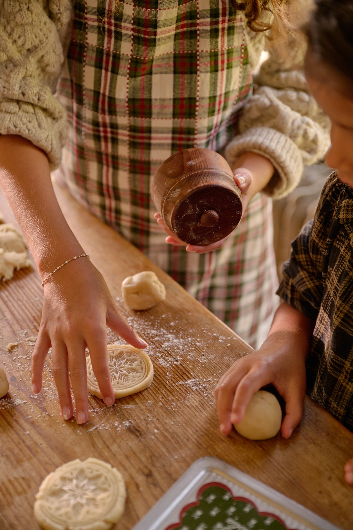 A cozy holiday scene showing a person and a child making gingerbread cookies at a wooden table. The person wears a beige knit sweater and a red, green, and white plaid apron, holding a dark wooden lidded container.