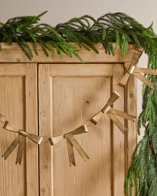 Gloria Brass Bow Garland shown hung on furniture with other greenery