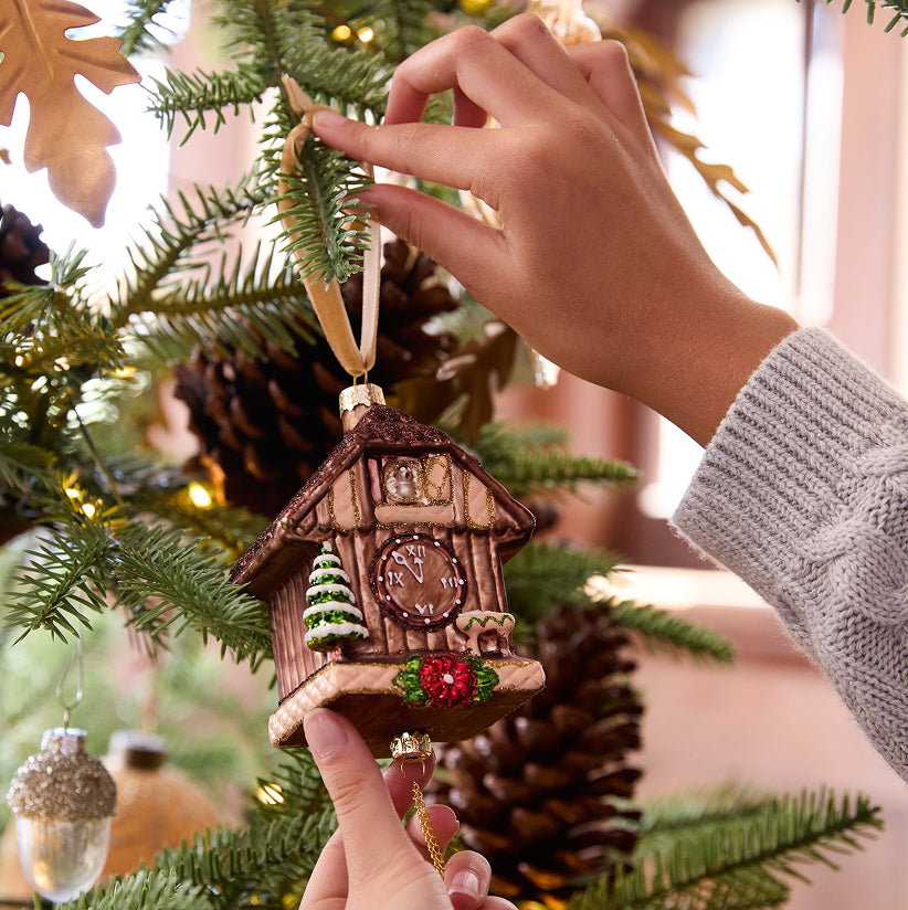 Hand placing a decorative Christmas cuckoo clock ornament on a tree.