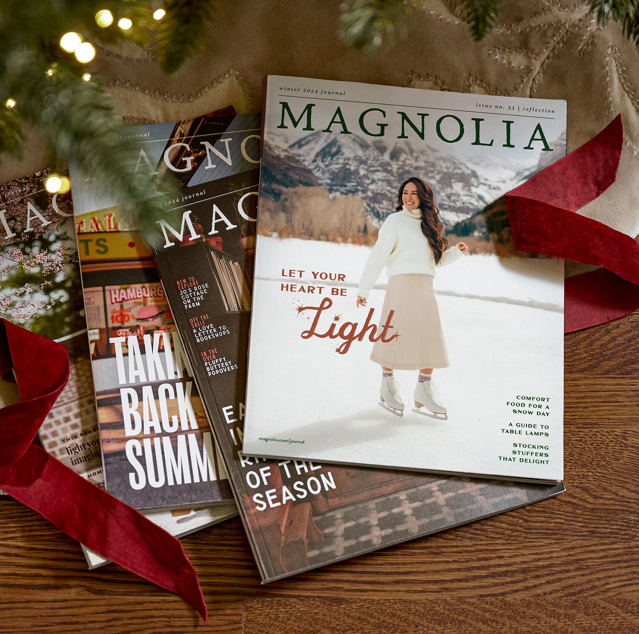 Magnolia Journal magazines on a wooden surface with decorative ribbons and greenery under a holiday tree.