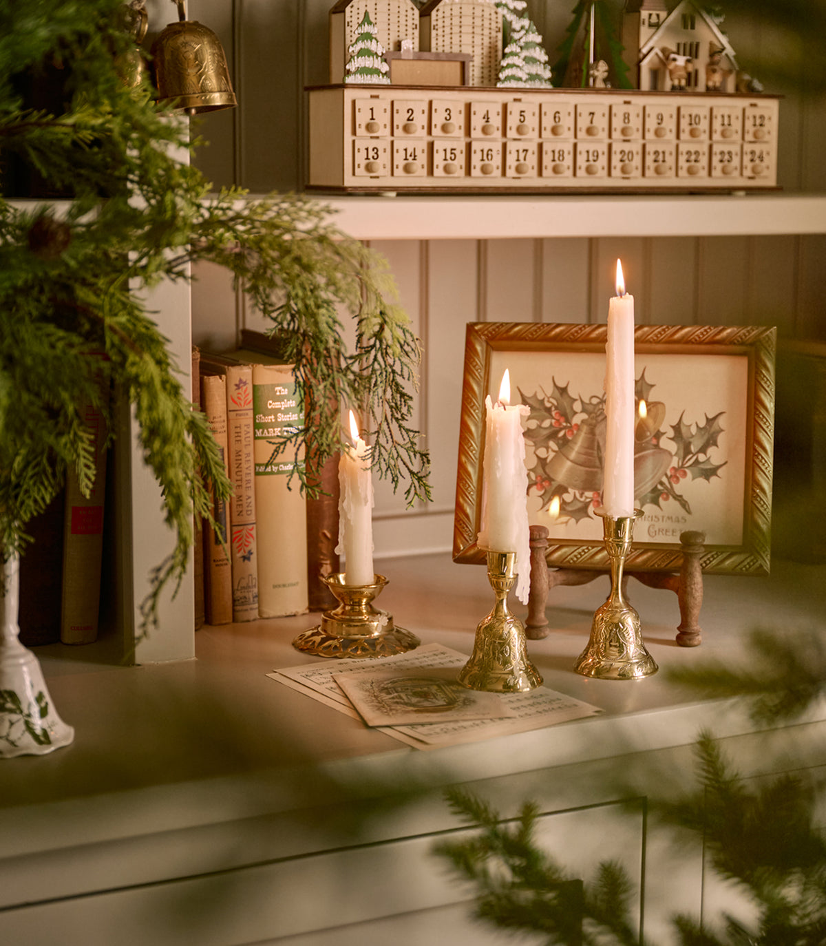 A cozy holiday tabletop scene with white taper candles in ornate gold candlesticks, a framed Christmas print, books, evergreen branches, and a shelf with decorative ornaments in the background.