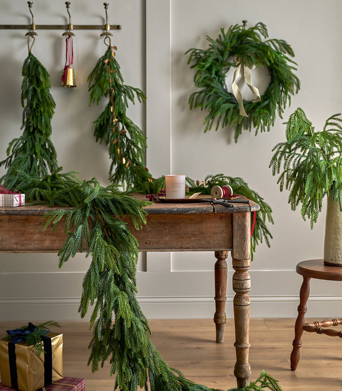 Green evergreen garlands and a matching wreath styled in a home setting, draped over a wooden table with holiday decor, gifts, and a vase on a side table against a neutral wall.