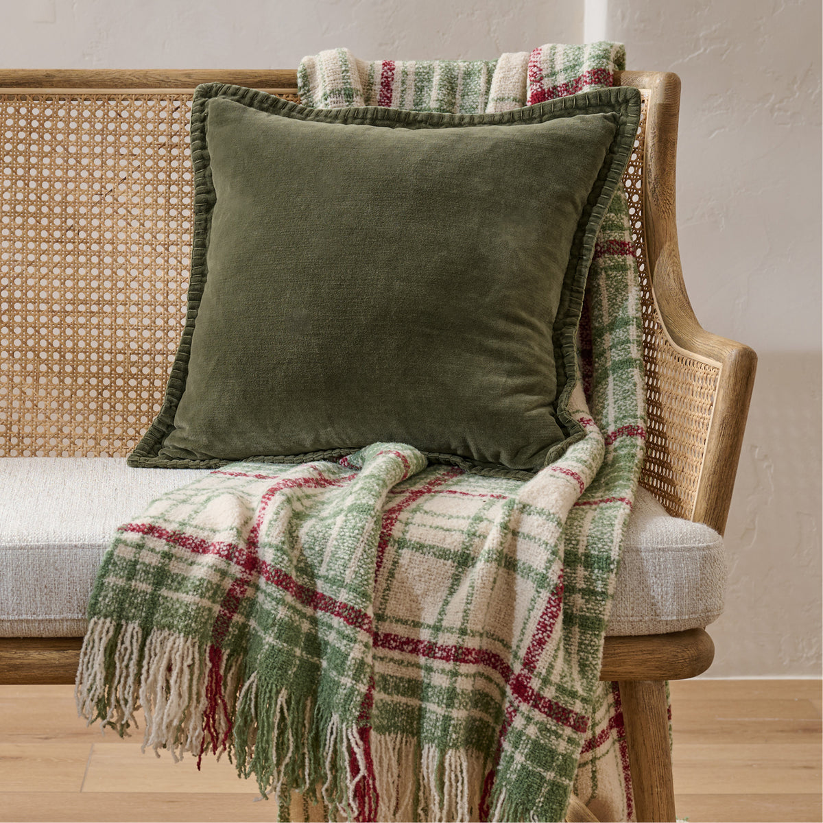 An olive green velvet throw pillow with a ruffled edge, styled on a wooden cane armchair draped with a green, red, and cream plaid fringed throw blanket.