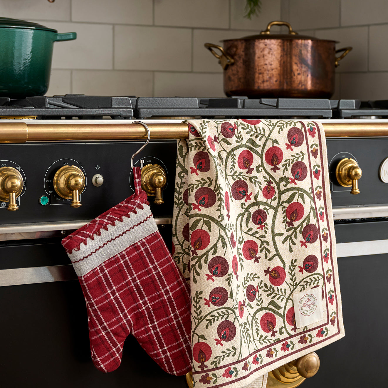 Red and white checkered oven mitt and patterned towel hanging on a stove with a copper pot in the background.