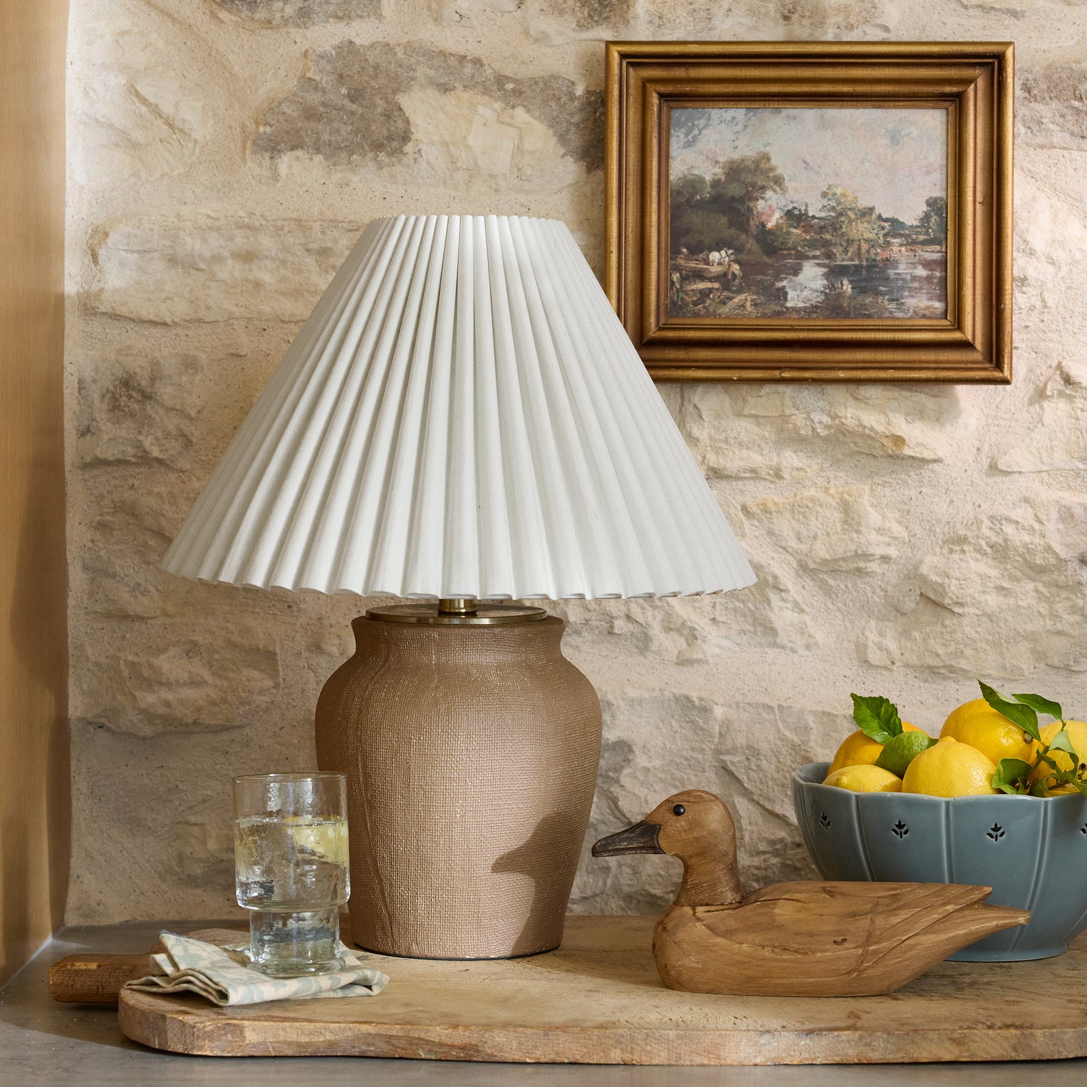 Lamp with a brown vase and white scalloped shade sitting on top of a wooden board.  Next to the lamp is a carved wooden duck and blue ceramic bowl full of lemons.