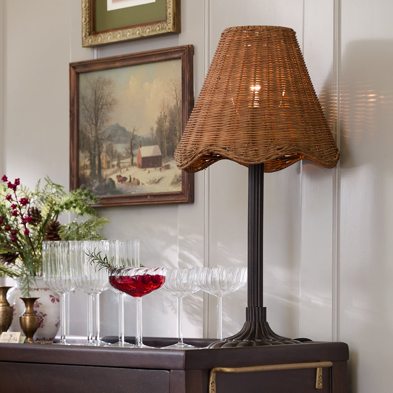 Wicker lamp on a sideboard with glasses and flowers in a room with framed artwork.