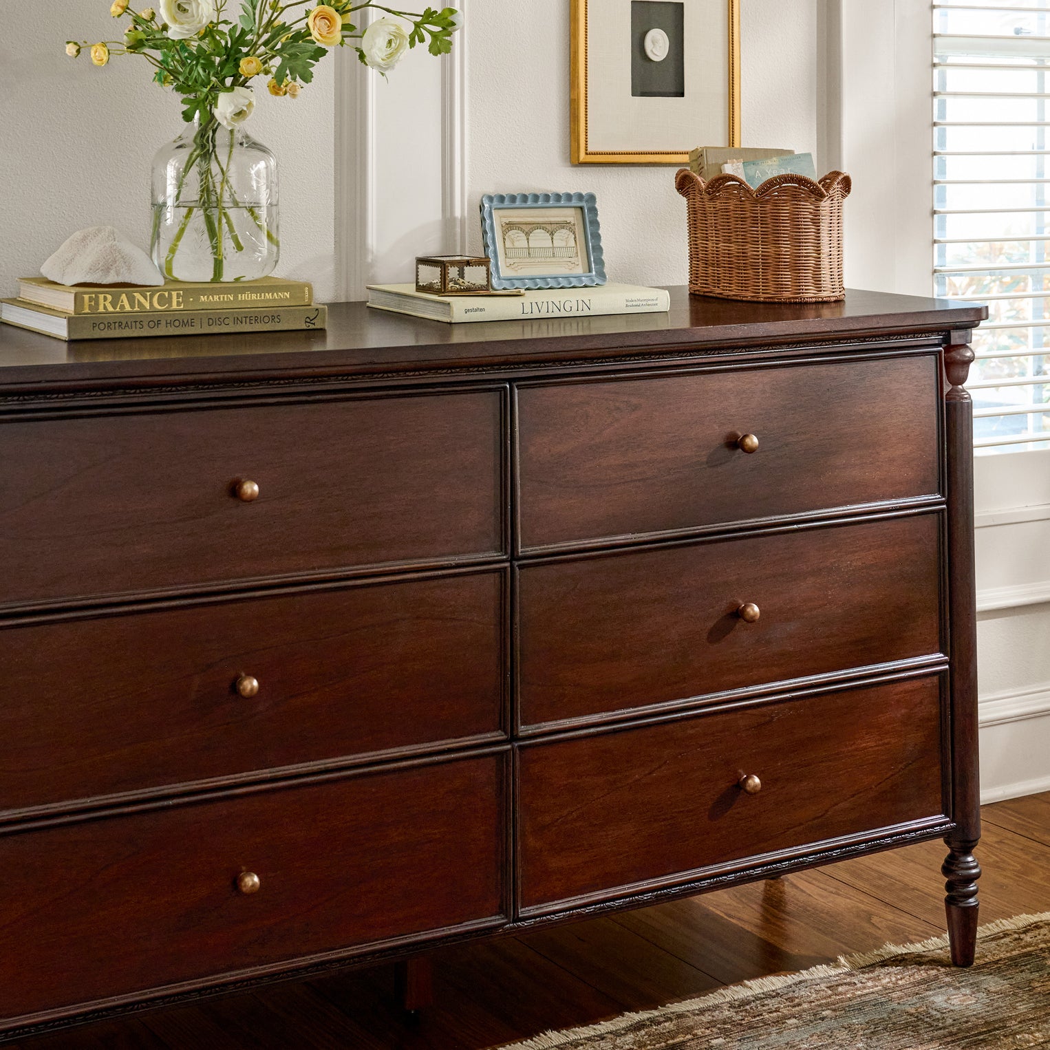 Brown six drawer dresser up against a wall.  On top of the dresser are stacked books, a blue frame, keepsake box, wicker basket and clear vase with yellow flowers.