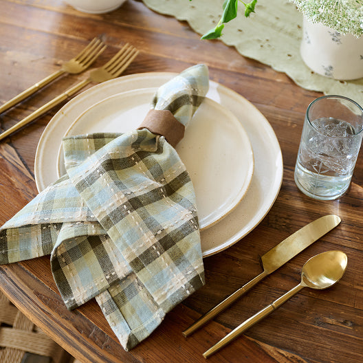 Plaid napkin on a cream lakelynn plate with gold cutlery and a glass of water on a wooden table.