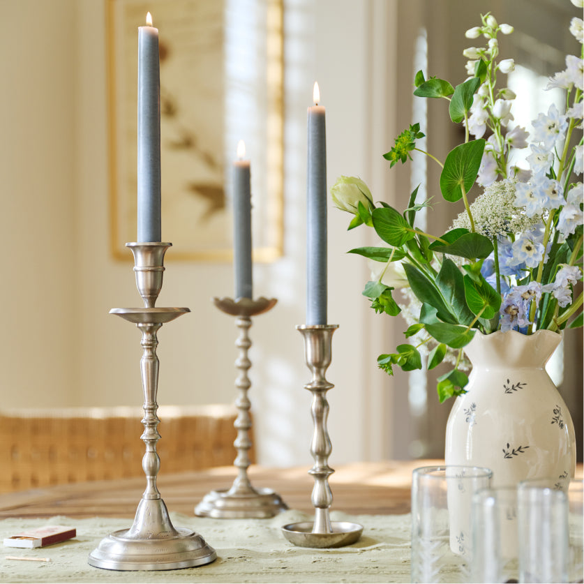 Decorative setup with silver candlesticks, gray candles, and a vase of flowers on a table.