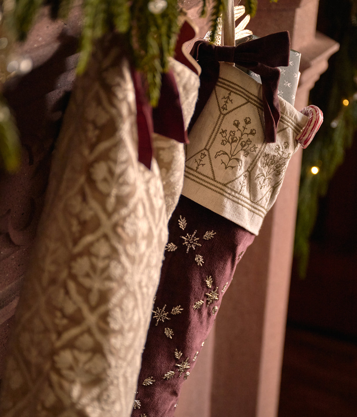 A close-up of two decorative Christmas stockings hanging side by side. One stocking is cream with a floral pattern and a dark burgundy bow, and the other is burgundy with cream embroidered snowflakes and a matching bow.