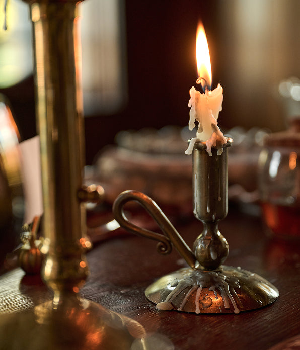 A close-up of a brass candlestick holder with a lit candle on a dark wooden table, with a blurred background and another brass object in the foreground.