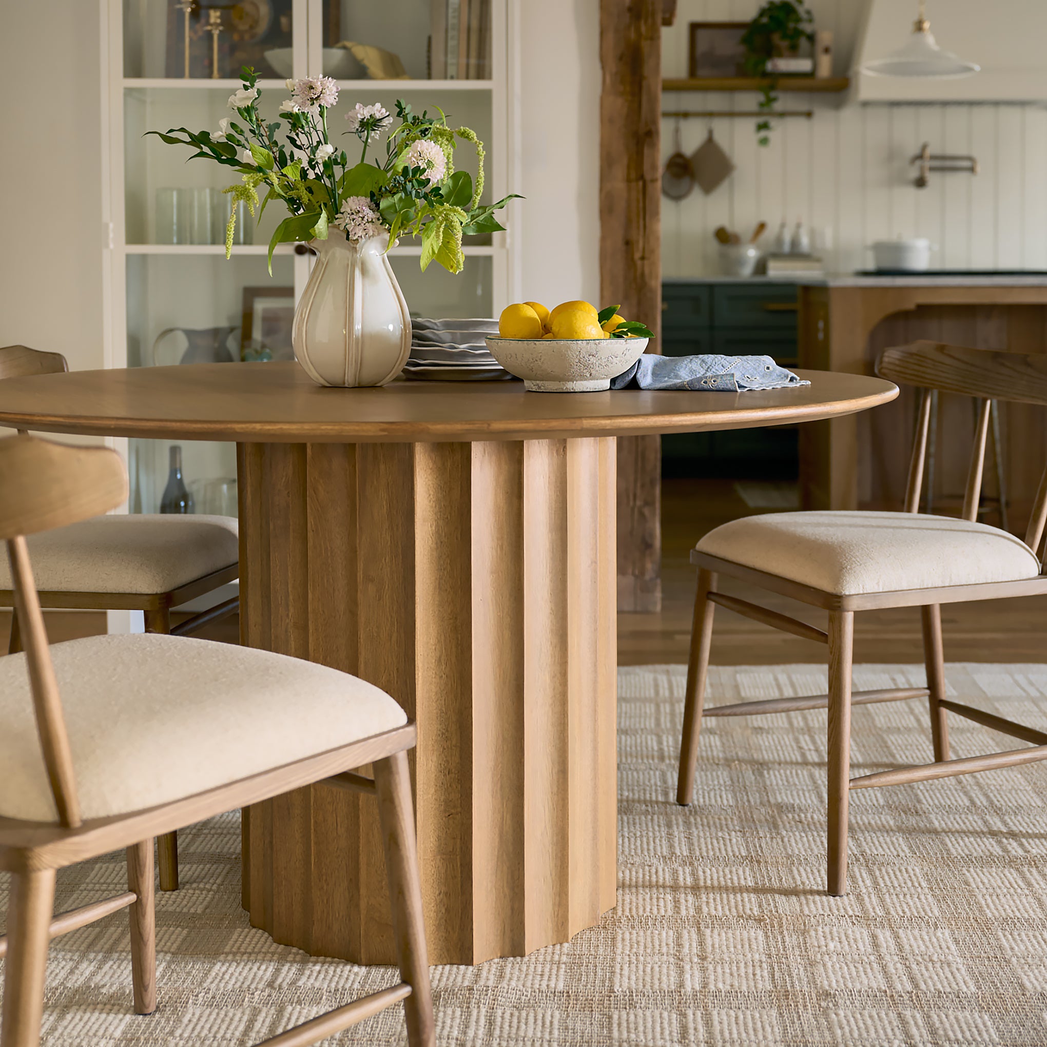 Dining room with wooden table and chairs, flowers, and kitchen in the background.