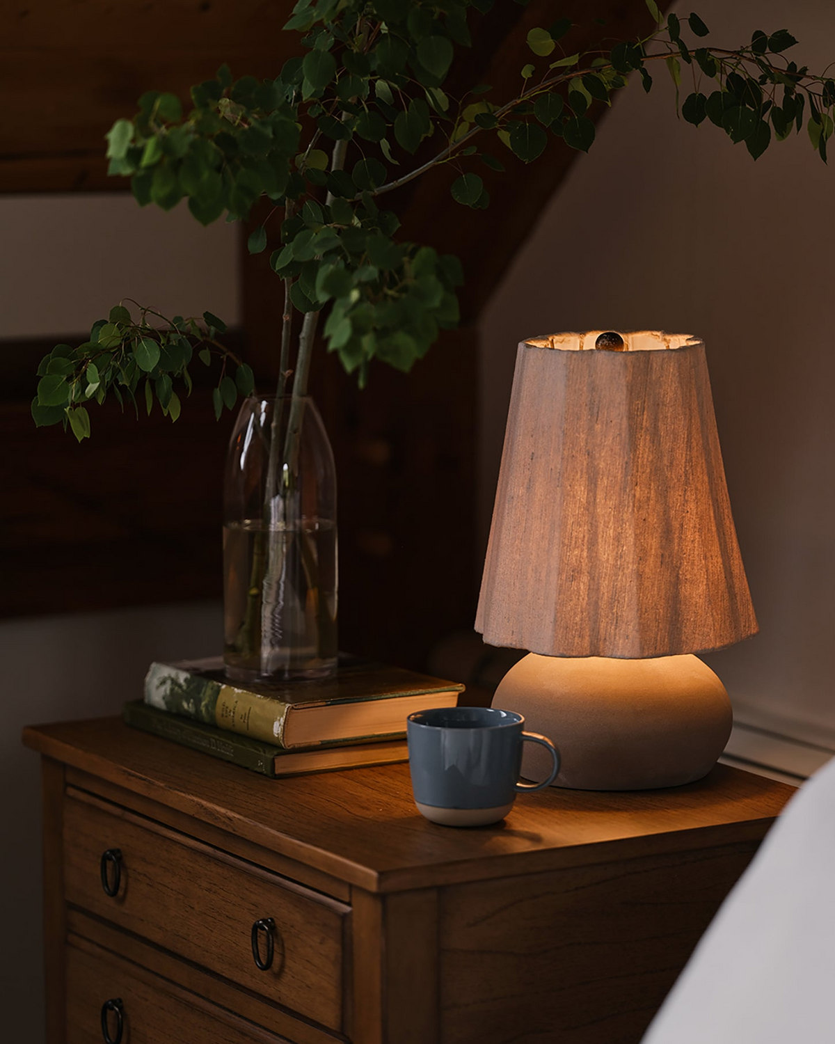 A warm lit table lamp with a beige shade and ceramic base sits on a wooden bedside table next to a blue mug, with books, a glass vase of greenery, and part of a bed visible in a cozy bedroom setting.
