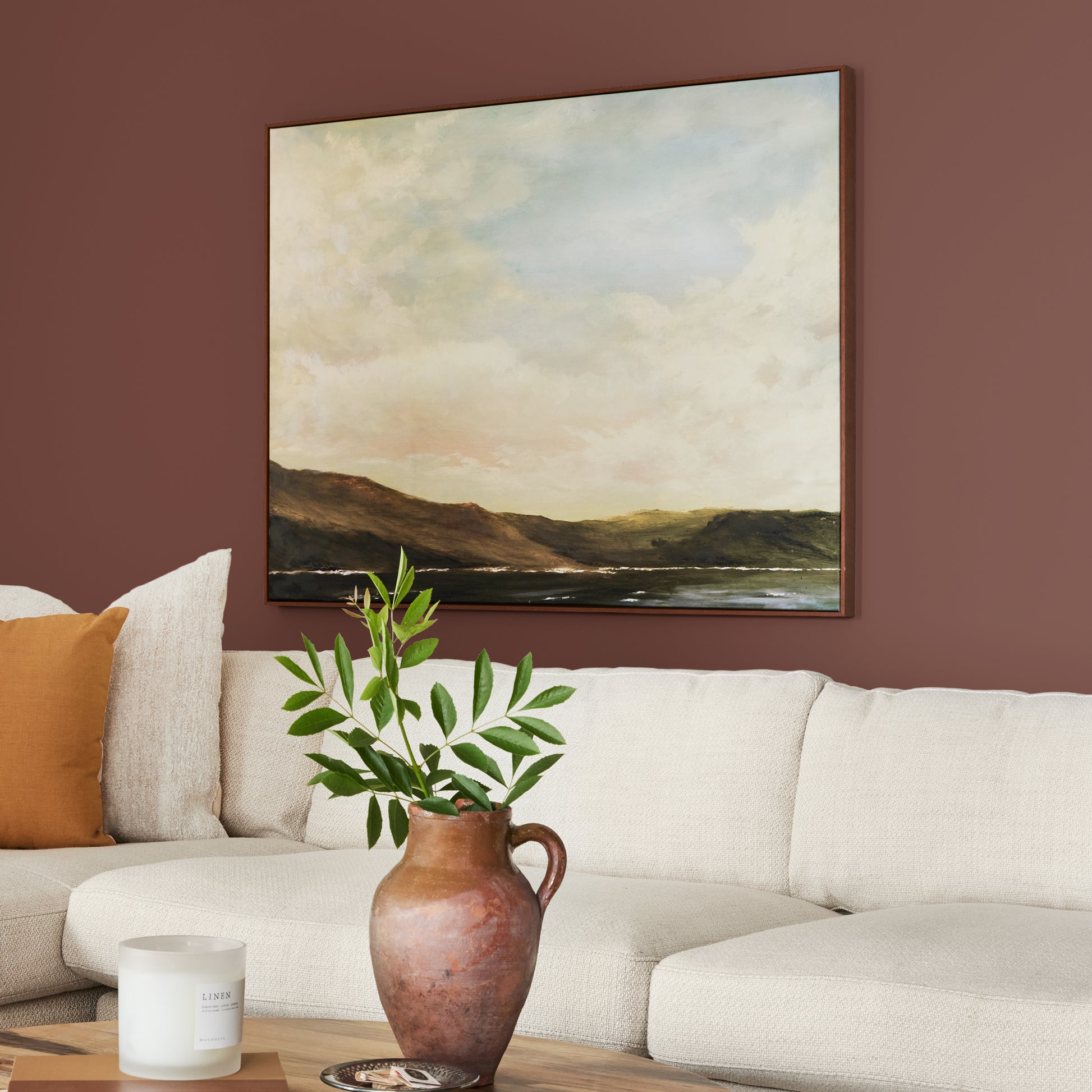 Mountain Currant paint featured in living room walls with large landscape picture above cream sectional and brown vase and greenery