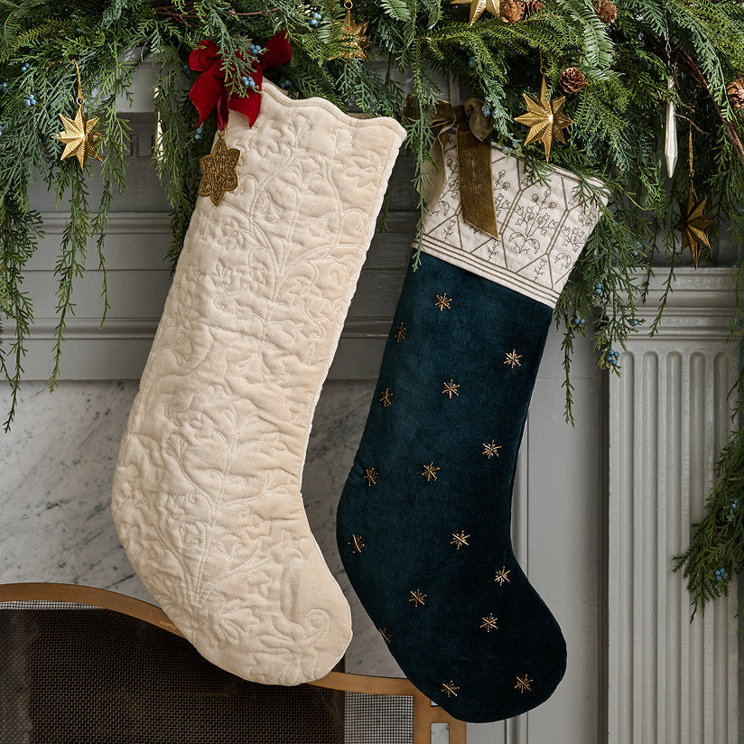 Two Christmas stockings, one white and one dark blue with gold stars, hanging on a fireplace mantel with greenery.