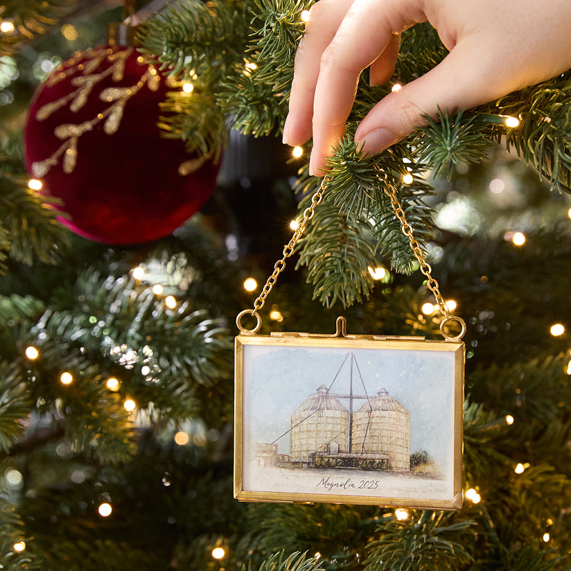Hand placing a decorative ornament with an illustration of the Silos on a Christmas tree.