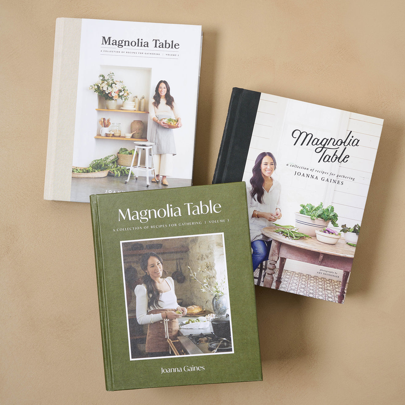 Three 'Magnolia Table' cookbooks by Joanna Gaines on a beige background.  shop cookbooks.