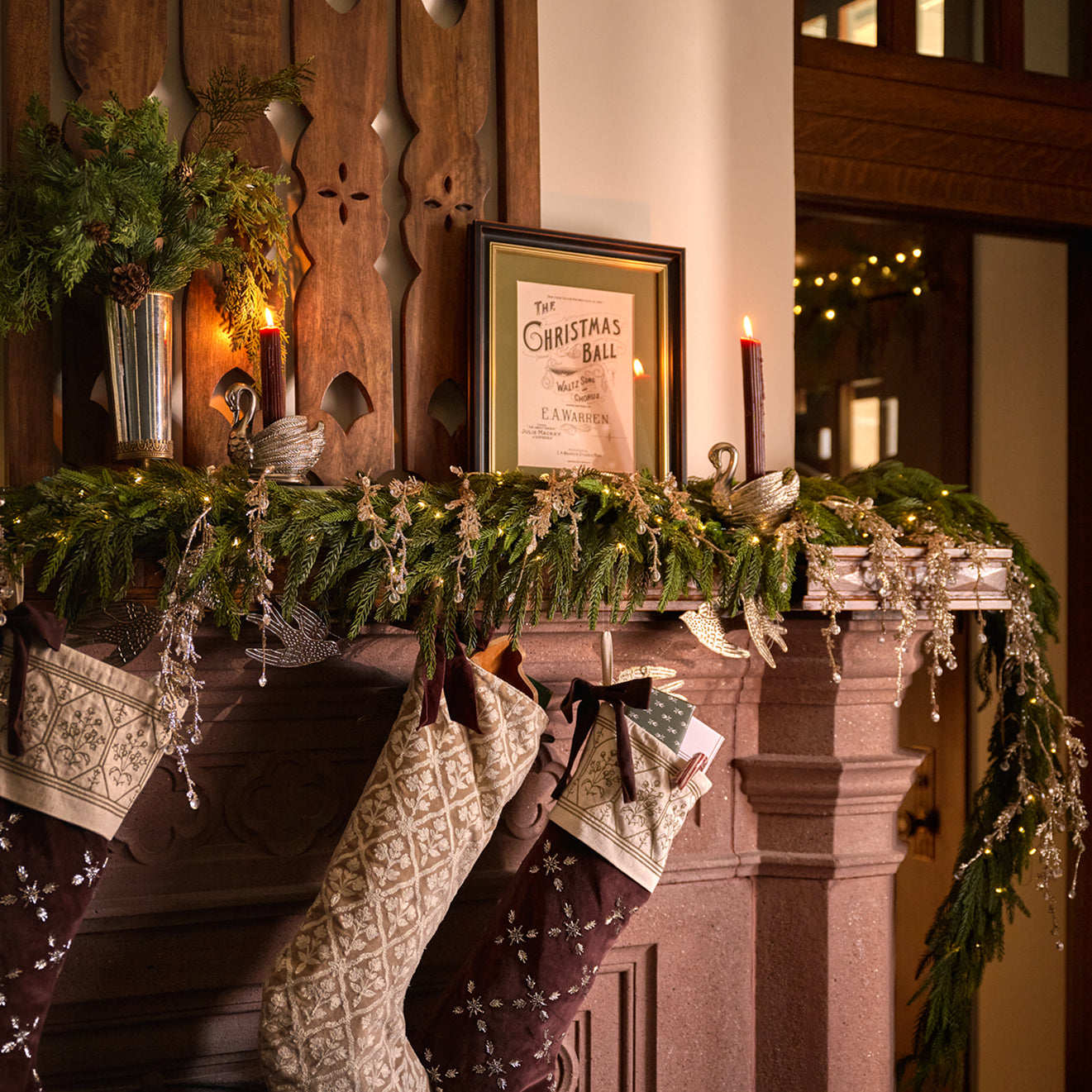 A cozy holiday fireplace scene with green garland draped across a stone mantel, lit candles, hanging patterned stockings, and a framed Christmas print against a dark wood paneled wall.