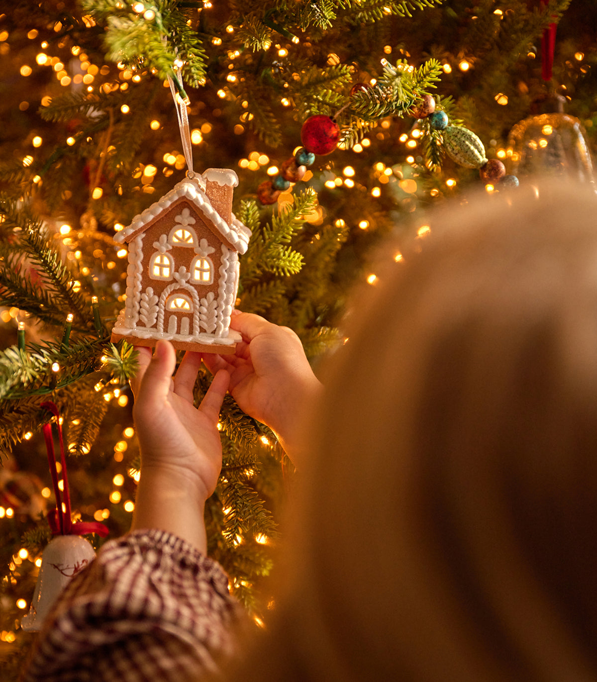 A person hangs a small gingerbread house Christmas ornament on a lit Christmas tree, with warm bokeh lights and green branches in the background.