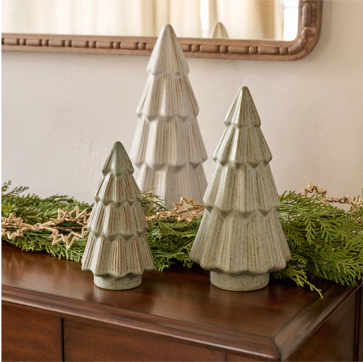 Three ceramic Christmas tree decorations on a wooden surface with greenery and a mirror in the background.